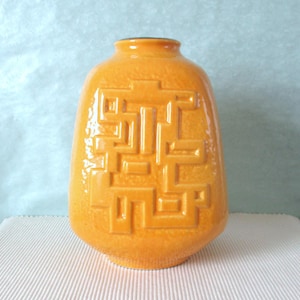 May include: An orange ceramic vase with a geometric pattern. The vase has a rounded shape and a narrow neck. The design is made up of interconnected rectangular shapes. The vase is on a white surface with a light blue background.