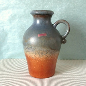 Può includere: Un vaso in ceramica con manico, caratterizzato da una sfumatura di colori. La parte superiore è blu scuro, che sfuma in un grigio-marrone maculato, e termina con una base rosso-arancio vibrante. È visibile una piccola etichetta.