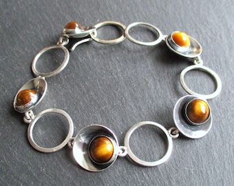 Puede incluir: Pulsera de plata con eslabones circulares y ovalados alternos. Cada eslabón ovalado contiene una piedra pulida, redonda, de color ámbar. La pulsera se muestra sobre una superficie gris oscuro, mostrando su diseño y materiales.