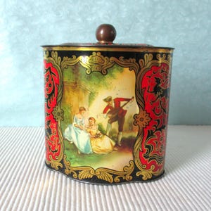 Op de afbeelding: Vintage blikken doos met een deksel en een bruine knop. De doos heeft een kleurrijke illustratie van een man die een snaarinstrument bespeelt voor twee vrouwen. De doos is versierd met rode, zwarte en gouden accenten.