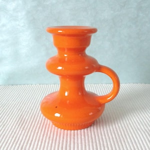 Könnte beinhalten: Ein leuchtend orangefarbener Kerzenhalter aus Glas mit rundem Sockel, bauchigem Mittelteil und ausgestelltem Oberteil. Er hat einen kleinen, gebogenen Griff an der Seite. Die glänzende Oberfläche reflektiert das Licht und hebt seine glatte, geschwungene Form hervor. Dieses Dekorationsstück eignet sich für die Inneneinrichtung.