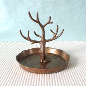 Könnte beinhalten: Ein bronzefarbener Schmuckbaum mit baumförmigem Design. Der Baum hat mehrere Äste zum Aufhängen von Schmuck und sitzt auf einer runden, blattförmigen Schale. Die Schale hat einen strukturierten, gewellten Rand.
