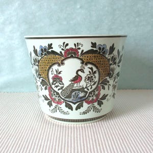 Op de afbeelding: Een witte keramische plantenbak met een decoratief pauwenontwerp. De plantenbak heeft een centraal medaillon met een pauwenillustratie, omgeven door bloemen en geometrische patronen in rood, zwart en goud. De rand en de basis hebben een donkerbruine rand.