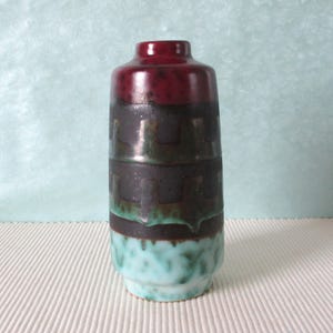 Peut inclure: Un vase en céramique avec un glaçage bordeaux, noir et turquoise. Le vase a un motif géométrique et une forme arrondie.