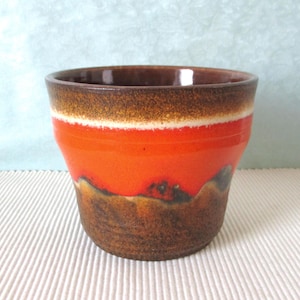 Könnte beinhalten: Eine Keramiktasse mit lebendigem Design. Sie hat einen dunkelbraunen Rand, einen weißen Streifen und einen leuchtend orangefarbenen Körper. Der Boden geht in ein dunkleres, strukturiertes Braun über. Diese dekorative Tasse eignet sich für Getränke oder als kleiner Pflanztopf.