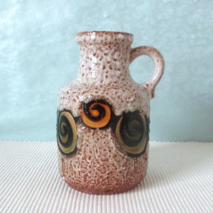May include: A white ceramic vase with a brown glaze and a textured surface. The vase has a handle and is decorated with a pattern of orange, green and black swirls.