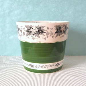 Può includere: Un vaso in ceramica verde con una fascia bianca con un motivo floreale bianco e nero. Il vaso ha un bordo ondulato e una finitura lucida.