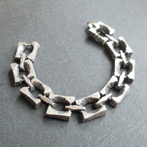 Puede incluir: Un brazalete plateado con un diseño de eslabones de cadena. Los eslabones son rectangulares con una apariencia texturizada y tosca. El brazalete se muestra sobre una superficie gris.