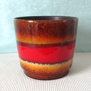Può includere: Un vaso di ceramica con finitura lucida. Il vaso presenta fasce orizzontali di colore, tra cui marrone, arancione e una vibrante striscia rossa. L'interno e il bordo sono di colore marrone scuro. Il vaso è progettato per uso interno o esterno.