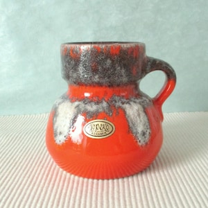 Könnte beinhalten: Eine kleine, orangefarbene Keramiktasse mit Henkel. Die Tasse hat einen strukturierten, gerippten Boden und ein Design aus weißen und grauen Akzenten. Der obere Rand ist dunkelbraun. Ein Aufkleber mit dem Text "Topeko Keramik" befindet sich auf der Vorderseite.