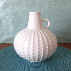 Può includere: Un vaso di ceramica bianca con corpo rotondo e superficie strutturata e puntinata. Il vaso ha un piccolo manico e un collo stretto. Si trova su un sottobicchiere turchese.