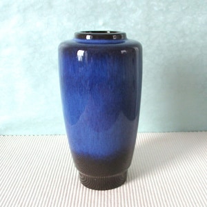 Könnte beinhalten: Eine hohe Keramikvase mit einem blauen Farbverlauf, der von einem dunkelbraunen Sockel zu einem tiefblauen Körper und einem dunkelbraunen Rand übergeht. Die Vase hat eine zylindrische Form mit leicht ausgestelltem Boden.
