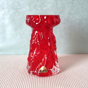 Peut inclure: Vase en verre rouge avec une surface texturée et irrégulière. Le vase a une base large et un col étroit. Le vase est fabriqué par la société 'Kosta Boda'.
