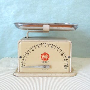 Può includere: Una bilancia da cucina vintage bianca con un vassoio argentato e un quadrante nero. Il quadrante è graduato in grammi e chilogrammi. La bilancia ha la scritta "Marke Rose ges. gesch. DRPA" stampata sul davanti.