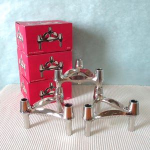 Può includere: Tre candelabri in metallo argentato, ciascuno con tre fori per candele. Dietro, tre scatole rosse con un disegno argentato del candelabro e la scritta "Made in Western Germany".