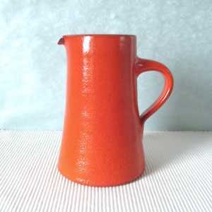 Può includere: Una brocca in ceramica color arancione brillante con una finitura superficiale testurizzata e leggermente granulosa. Il recipiente presenta un corpo cilindrico che si restringe leggermente verso l'alto, un piccolo beccuccio e un manico curvo.