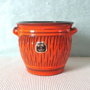 Peut inclure: Un pot de fleurs en céramique orange vif avec un motif de rainures verticales texturées. Le pot a deux petites poignées et un bord foncé. Une petite étiquette avec du texte est apposée sur le devant. Une pièce décorative.
