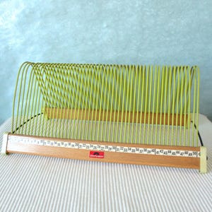 Könnte beinhalten: Ein limettengrüner Schallplattenständer aus Draht mit einem Holzsockel, der eine nummerierte Skala von 1 bis 50 aufweist. Auf der Vorderseite des Holzsockels befindet sich ein rotes Polydor-Logo. Der Ständer hat mehrere gebogene Drahtfächer.