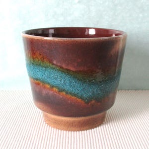 Op de afbeelding: Een keramische beker met een uniek ontwerp. De beker heeft een rijke, donkerbruine buitenkant met een levendige turquoise band. De basis is lichter bruin, wat een opvallend contrast creëert. De beker is ongeveer 9 cm hoog.