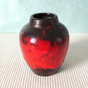 Könnte beinhalten: Eine Keramikvase mit rundem Körper und schmalem Hals. Die Vase weist einen auffälligen Farbverlauf auf, der von einem tiefen, dunklen Braun oben zu einem leuchtenden Rot am Boden übergeht. Der Boden hat ein strukturiertes, geriffeltes Design.