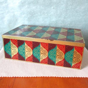 Può includere: Una scatola rettangolare in metallo con un motivo geometrico ripetitivo di forme a goccia color verde acqua, rosso e viola scuro con dettagli a volute dorate. Il coperchio presenta un bordo dorato e riprende il design colorato del corpo.