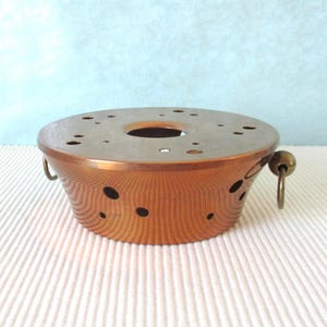 Puede incluir: Un soporte redondo de metal color cobre para quemador de fondue, con una parte superior plana, una abertura circular central y múltiples orificios de ventilación. Los lados son cónicos con recortes circulares adicionales y dos asas de anillo.
