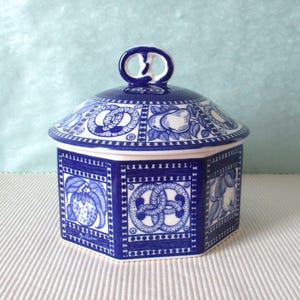 Peut inclure: Un pot hexagonal en céramique avec un couvercle présentant un motif bleu et blanc. Le design comprend des illustrations de divers fruits et des motifs de nœuds géométriques dans des panneaux rectangulaires. Le couvercle a une anse.