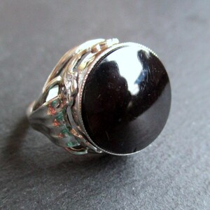 Può includere: Un anello d'argento con un designo ornato e una grande pietra preziosa nera.
