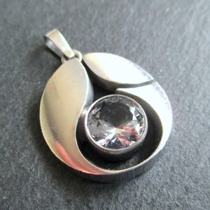 Puede incluir: Un colgante circular de metal plateado con acabado pulido y una piedra preciosa redonda y transparente facetada en el centro. El colgante tiene un diseño de marco curvo y liso con una anilla integrada en la parte superior para una cadena.