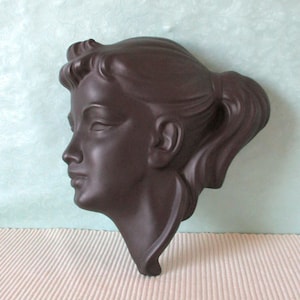 Könnte beinhalten: Eine dunkelbraune Wandskulptur, die das Profil einer Frau darstellt. Die Skulptur zeigt detaillierte Gesichtszüge, darunter geschlossene Augen, eine definierte Nase und einen Pferdeschwanz. Das Stück ist wahrscheinlich aus Keramik oder einem ähnlichen Material gefertigt.