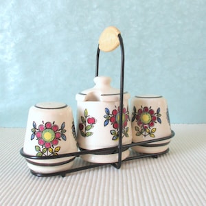 Può includere: Un set di tre saliere e pepiere in ceramica con un design floreale. Le saliere sono bianche con fiori rossi, gialli e blu. Le saliere sono su un supporto in metallo nero con un manico.