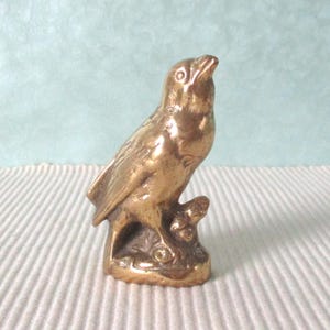 Puede incluir: Pequeña figura de pájaro de latón, posada en una rama. El pájaro mira hacia arriba con el pico abierto. La figura tiene una superficie detallada y texturizada, sobre un fondo azul claro y blanco.
