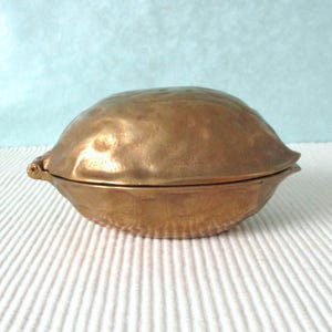 Könnte beinhalten: Eine Vintage-Schmuckschatulle in Messingoptik in Form einer Walnuss. Die Box hat einen Klappdeckel und eine strukturierte Oberfläche mit einem detaillierten Design, das die natürliche Form einer Walnuss nachahmt. Die Box steht vor einem hellblauen Hintergrund.