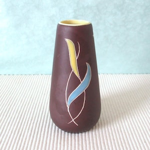 Könnte beinhalten: Eine kleine, braune Keramikvase mit einem gelben und blauen abstrakten Design. Die Vase hat einen abgerundeten Boden und eine schmale Öffnung.
