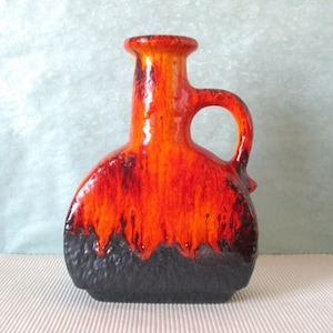 Pode incluir: Um vaso de cerâmica com uma pega, apresentando um gradiente de cores marcante. O topo é laranja vibrante, passando para vermelho e depois para uma base preta texturizada. O vaso tem um corpo arredondado e um pescoço cilíndrico.