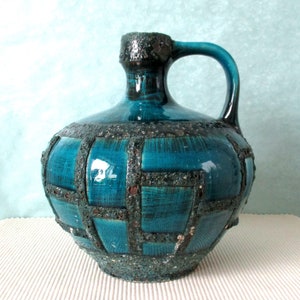 May include: A teal ceramic pitcher with a dark handle and textured rim. The body features a grid-like pattern in a darker shade, creating a striking contrast. The jug has a rounded shape and a sturdy handle, perfect for serving beverages.