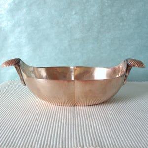 Peut inclure: Un bol en cuivre poli avec un corps lisse et incurvé et deux poignées en forme de coquillages sur les côtés opposés. Le bol présente une ouverture large et une base arrondie, reflétant la lumière sur sa surface métallique aux tons cuivrés.