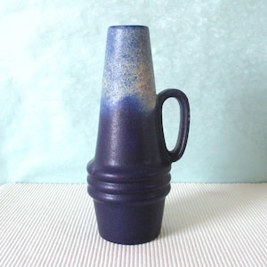 Może przedstawiać: Ciemnoniebieski ceramiczny wazon z uchwytem. Wazon ma rozszerzoną podstawę i cylindryczny korpus, który zwęża się ku górze. Górna część jest jaśniejsza niebieska z nakrapianymi detalami. Uchwyt ma prosty kształt pętli.