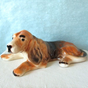 Könnte beinhalten: Eine Keramikfigur eines braunen und schwarzen Basset-Hundes, der auf dem Bauch liegt. Der Hund hat ein langes, hängendes Gesicht und schlaffe Ohren.