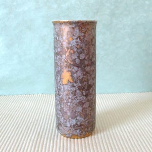 Könnte beinhalten: Eine hohe, zylindrische Vase mit einem gesprenkelten Muster in Braun-, Lila- und Goldtönen. Die Vase hat einen goldenen Rand und Boden sowie einen goldfarbenen, blattartigen Akzent. Der Hintergrund ist hellblau.