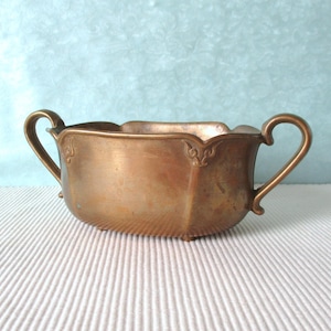 Pode incluir: Uma tigela de latão vintage com duas alças curvas. A tigela tem uma borda ondulada e uma base texturizada e canelada. Detalhes ornamentais são visíveis perto das alças. A tigela é de uma cor dourada antiga quente.