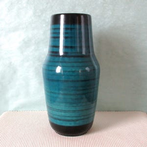 Könnte beinhalten: Eine türkisfarbene Keramikvase mit horizontalen schwarzen Streifen. Die Vase hat einen dunkelbraunen Rand und Boden. Die glänzende Oberfläche reflektiert das Licht und hebt die Farbgebung Türkis und Schwarz hervor. Diese dekorative Vase eignet sich für die Inneneinrichtung.