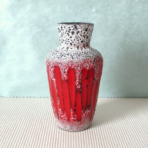 Può includere: Un vaso in ceramica caratterizzato da un corpo rosso vibrante con motivi a goccia verticali. Il collo e il bordo superiore presentano una finitura in smalto bianco testurizzato con macchie nere, creando un forte contrasto con la base rossa lucida.