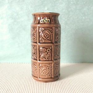 Può includere: Un vaso in ceramica marrone con un motivo geometrico di foglie e fiori. Il vaso ha un'etichetta dorata con il testo "Bav".