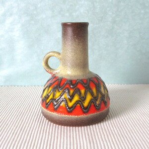 Peut inclure: Un petit vase en céramique brun avec une anse. Le vase a un motif géométrique jaune, orange et noir.