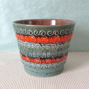 Könnte beinhalten: Ein Keramik-Blumentopf mit strukturierter Oberfläche und farbenfrohem Design. Der Topf hat Bänder in Orange, Grün und Blau mit dekorativen Mustern. Der Innenraum ist dunkelbraun.