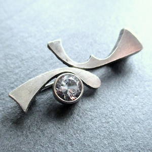 Puede incluir: Un broche abstracto de plata con un cristal transparente en el centro.