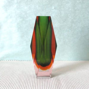 Peut inclure: Un vase en verre à facettes, haut, présentant un panneau central vert profond flanqué de côtés orange vif. La base est transparente et la structure se rétrécit vers le haut avec des bords nets et anguleux, créant une silhouette géométrique.