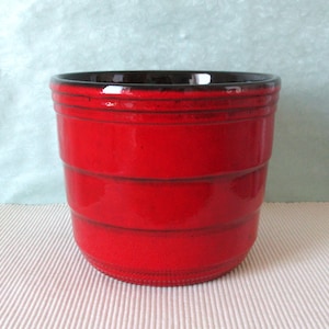 Puede incluir: Una maceta de cerámica roja brillante con acabado brillante. La maceta presenta detalles horizontales acanalados y un interior marrón oscuro. Adecuada para uso en interiores o exteriores, es un elemento decorativo.