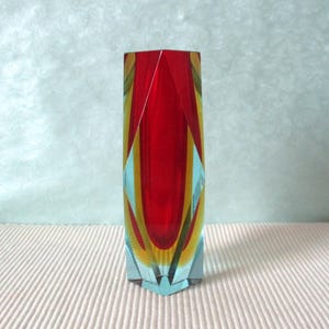 Könnte beinhalten: Eine hohe, facettierte Glasvase mit einem leuchtend roten Mittelteil, der von vertikalen Schichten aus gelbem und klarem Glas flankiert wird. Das geometrische Design erzeugt scharfe, kantige Formen und einen prismatischen Effekt.